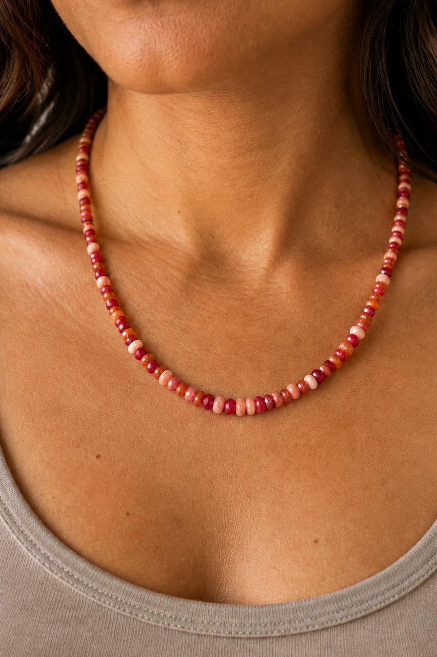 A close-up of a woman wearing a beaded necklace made of small round beads in shades of red, pink, and orange, paired with a gray ribbed tank top.