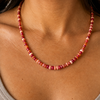 A close-up of a woman wearing a beaded necklace made of small round beads in shades of red, pink, and orange, paired with a gray ribbed tank top.